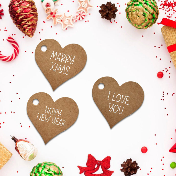 Drie hartvormige kartonnen labels met teksten 'MARRY XMAS', 'HAPPY NEW YEAR' en 'I LOVE YOU' op een kerstdecoratie achtergrond.